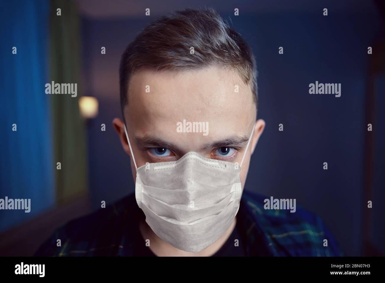 Sconvolto in isolamento domestico, un giovane uomo caucasico. Il look pazzo di un maschio bianco che indossa una maschera protettiva al coperto durante il periodo di coro Foto Stock