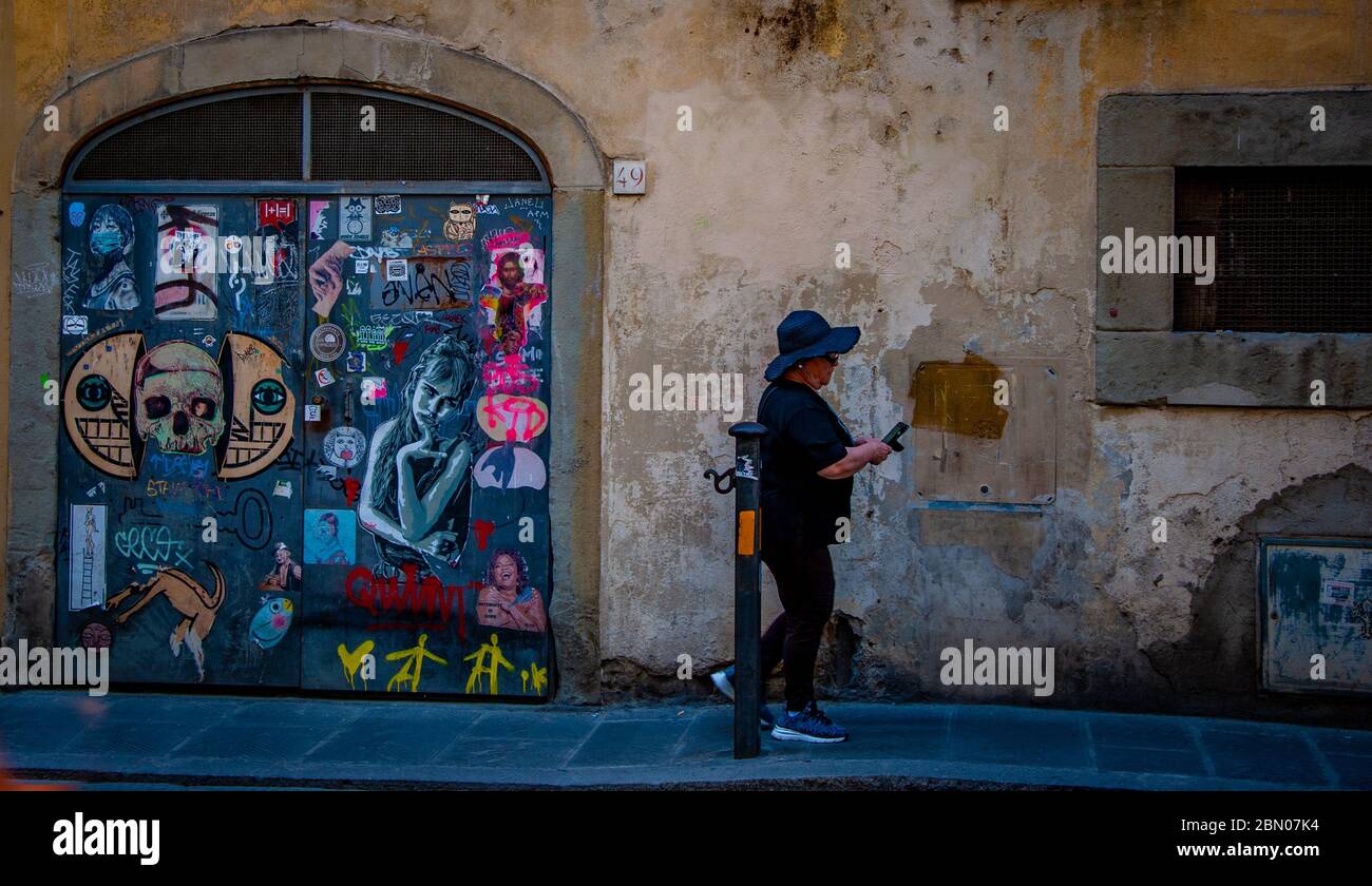Una donna di mezza età non identificabile in scarpe da running usa un Telefono cellulare in una rustica strada italiana che passa un arco porta coperta di arte di strada Foto Stock