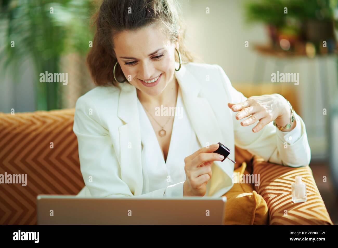 donna sorridente ed elegante in stile di mezza età con blusa e giacca bianca con chiodo lucido per laptop nel moderno soggiorno in giornata di sole. Foto Stock