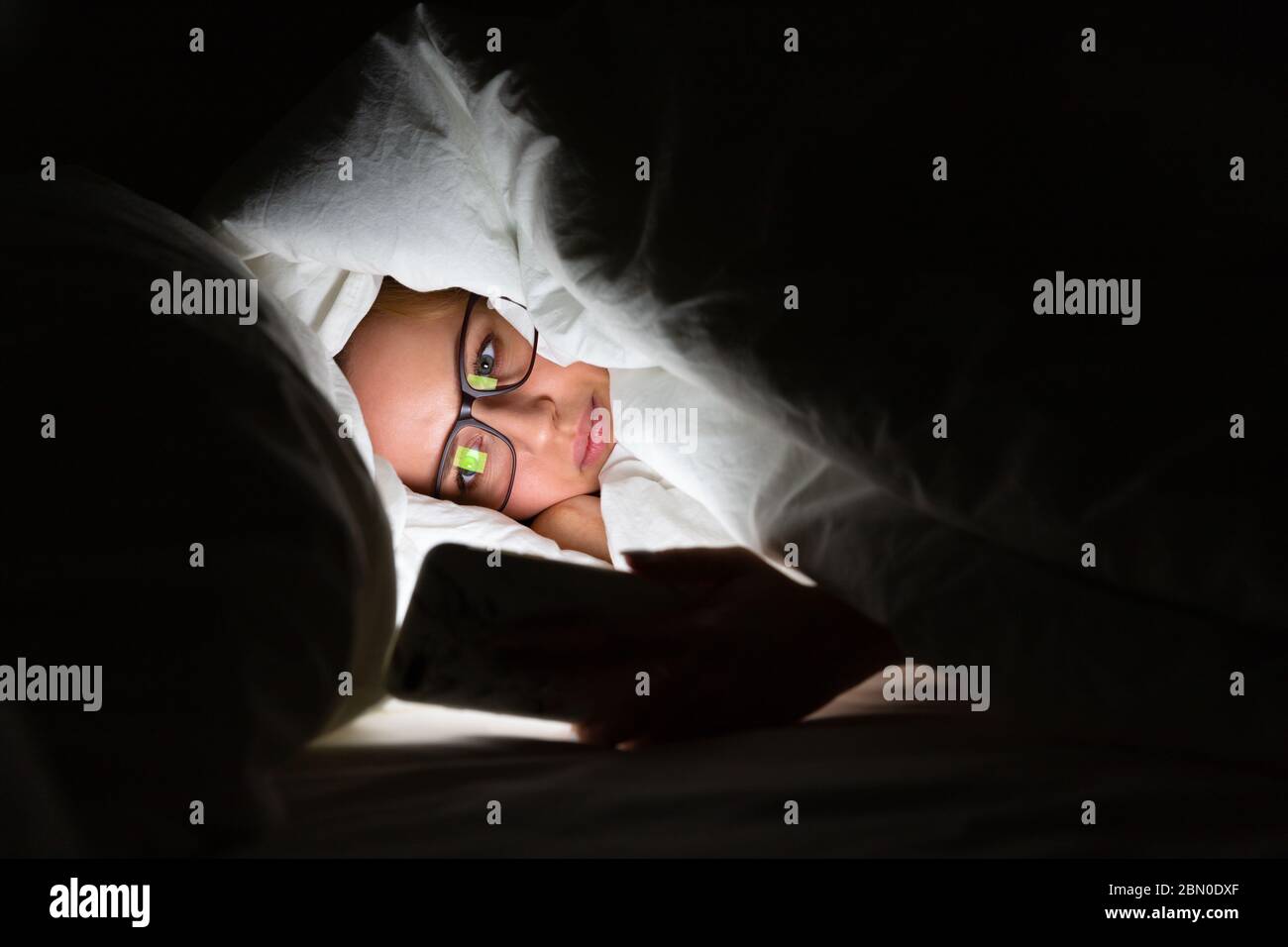 Donna in occhiali che si trova a letto sotto la coperta utilizzando smartphone a tarda notte, non può dormire, addict notizie su Covid-19 o scorrimento social network Foto Stock