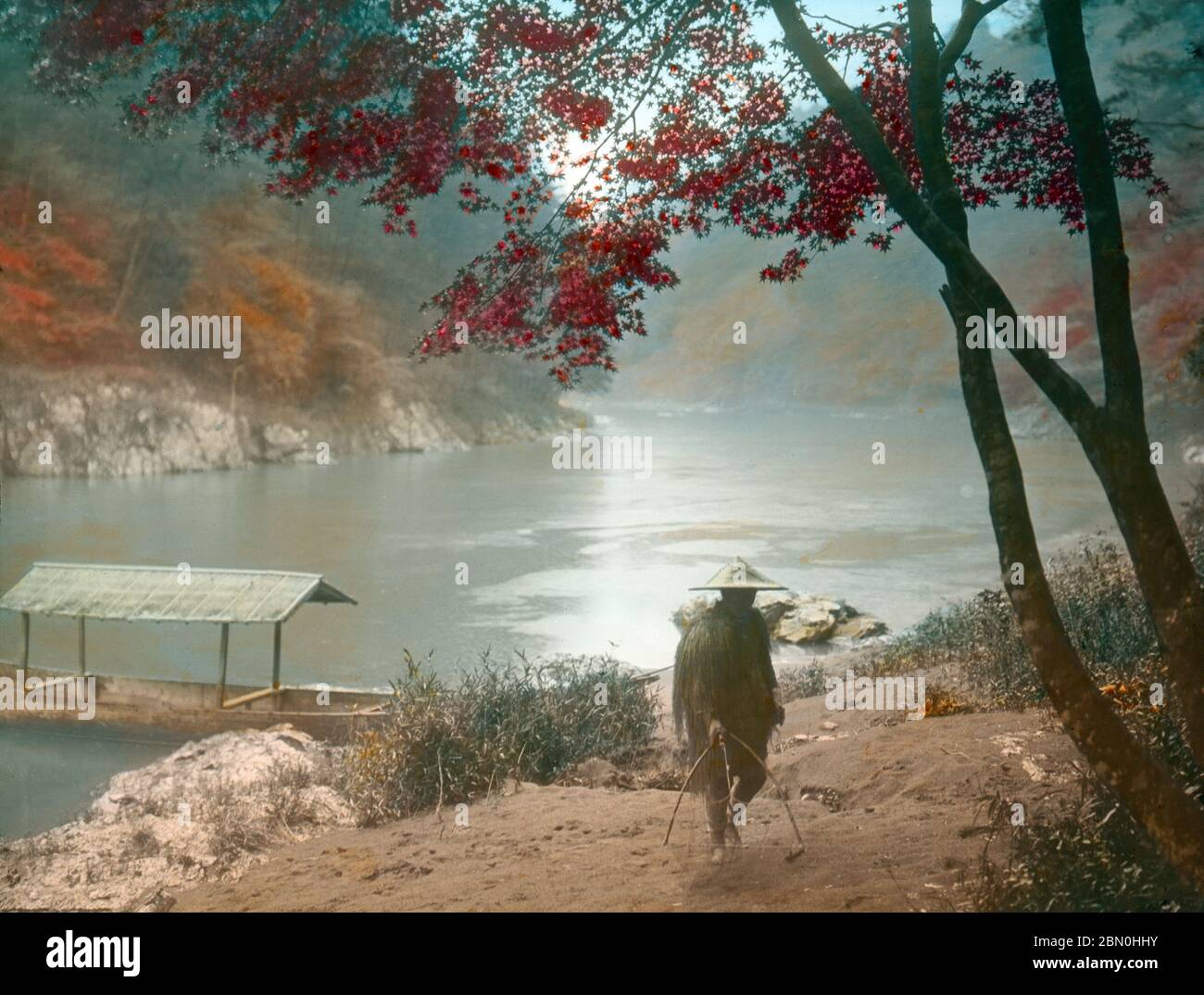 [ 1900 Giappone - Autunno ad Arashiyama, Kyoto ] - un uomo che indossa un impermeabile (mino) e un cappello conico (菅笠, sugegasa) trasporta una rete di pesca durante l'autunno al fiume Hozugawa ad Arashiyama, Kyoto. Sul retro si può vedere una barca a fondo piatto. Per secoli il fiume Hozugawa è stato utilizzato per trasportare merci come riso, orzo, grano, carbone e legname. Ma dal 1895 (Meiji 28), il viaggio di 2 ore lungo il fiume divenne una popolare attrazione turistica (Hozugawa Kudari). vetrino vintage del xix secolo. Foto Stock