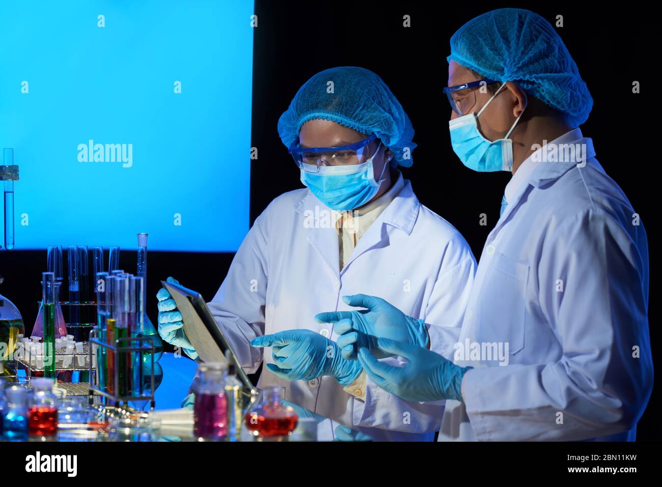 Tecnici di laboratorio in maschere mediche che discutono i dettagli della ricerca sul computer tablet prima di miscelare i reagenti Foto Stock