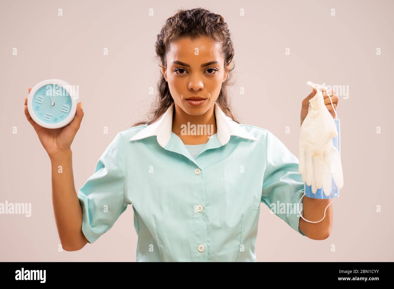 Ritratto di infermiere serio che sta tenendo indumenti da lavoro protettivi e orologio che mostra da cinque a dodici volte. Foto Stock