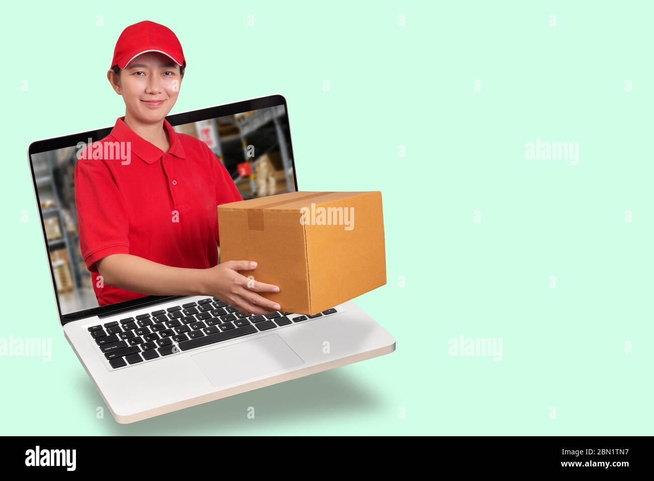 Donna asiatica consegna in camicia rossa con sfondo sfocato magazzino, tenendo ordine merci in pacco fuori dal computer portatile isolato Foto Stock