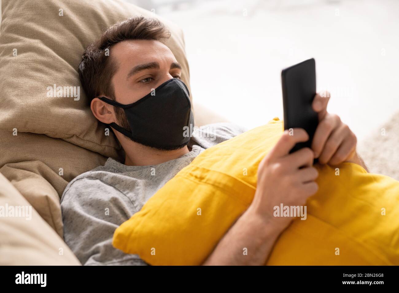 Giovane uomo serio in maschera protettiva sdraiato con cuscino sul divano e utilizzando smartphone mentre legge le notizie sul coronavirus Foto Stock