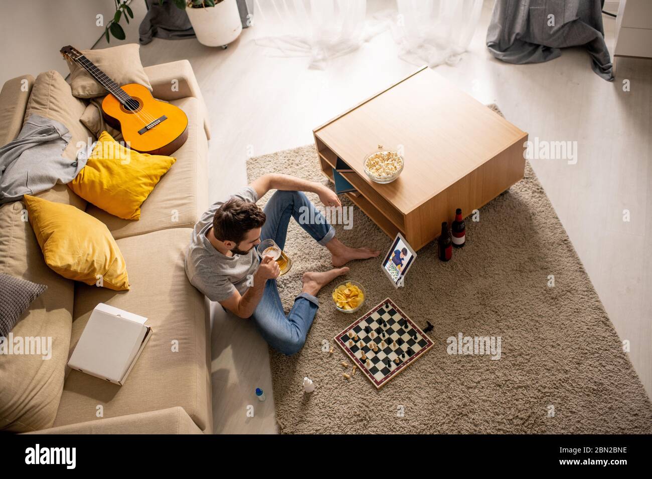 Sopra la vista del giovane uomo seduto in salotto e bere birra mentre trascorrere il tempo con l'amico online durante la quarantena Foto Stock