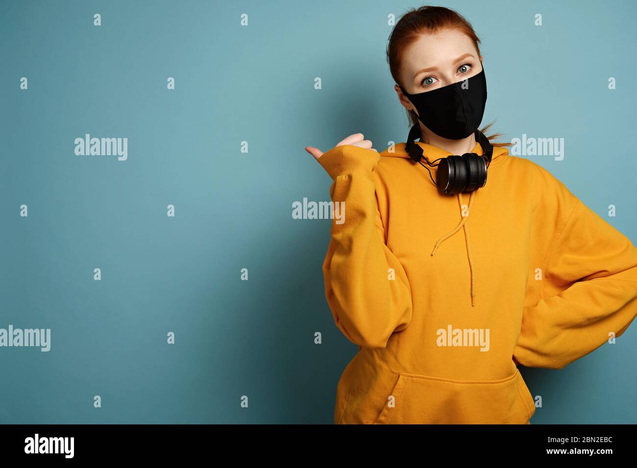 La ragazza in una felpa con cappuccio gialla e maschera protettiva nera si trova su uno sfondo blu e punta il pollice verso il lato Foto Stock