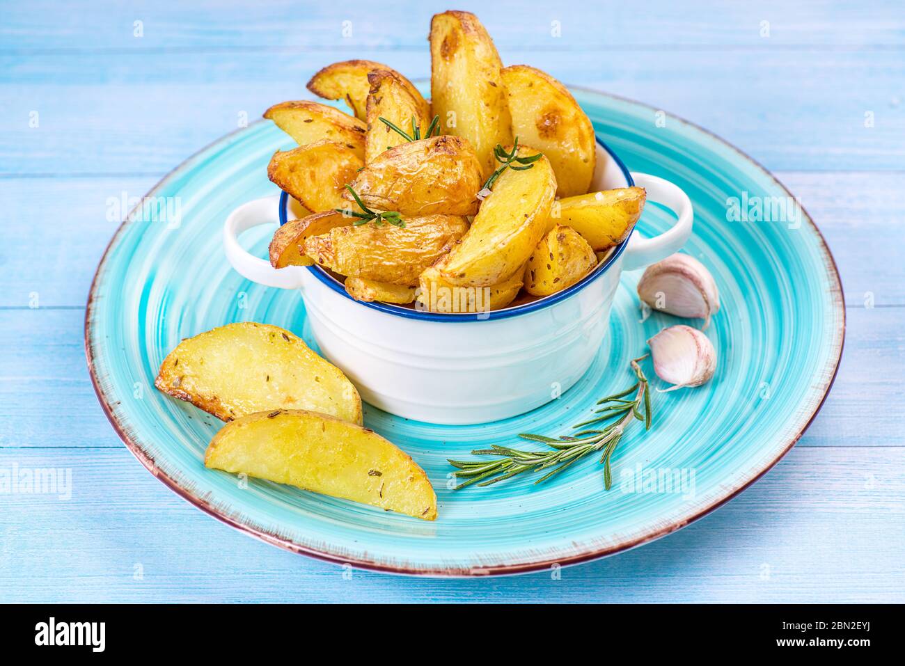 Patate fritte incastrano le patate in un piatto blu su sfondo di legno blu con aglio e un ramo di razmarin. Foto Stock