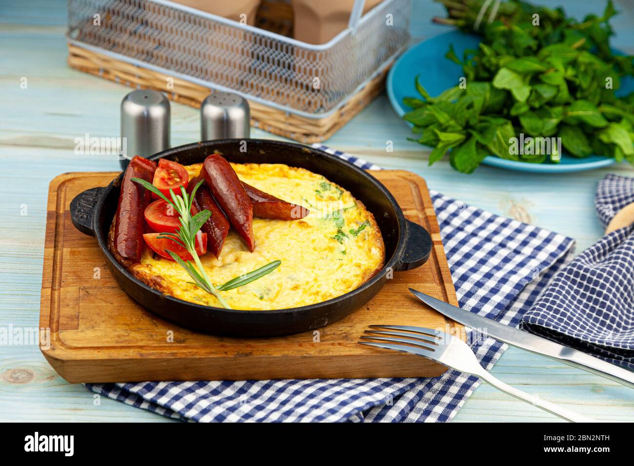 Omelette con salsiccia e pomodori in una padella sul tavolo. Colazione appetitosa. Un rametto di rosmarino, una spezia di legno. Caccia salsicce. Cibo gustoso fritto Foto Stock