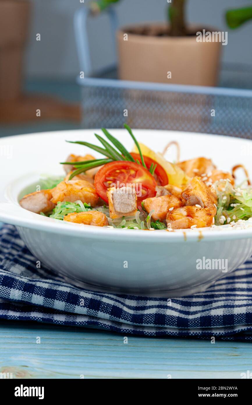 Tagliatelle di riso con salmone e spinaci. Pranzo in un piatto bianco sul tavolo. Pomodori, limone, pesce, rosmarino, semi di soi e sesamo. Cibo ancora vita Foto Stock