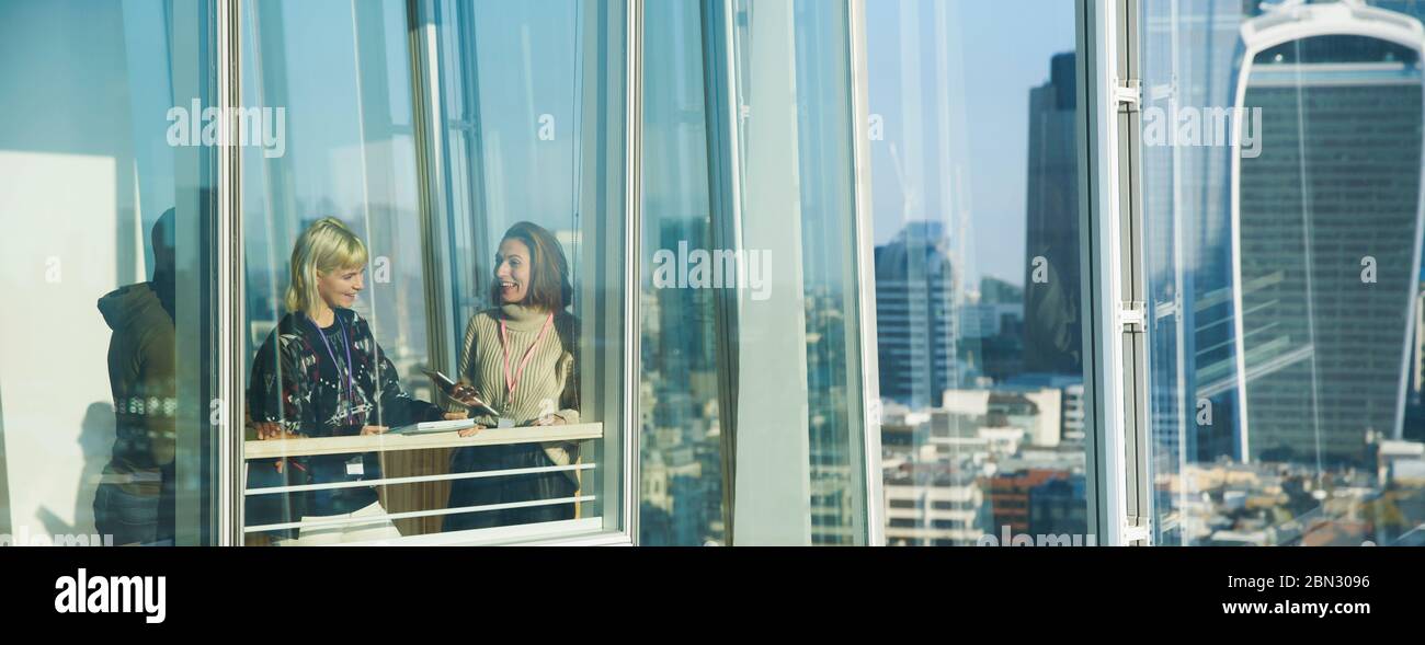 Donne d'affari che parlano alla soleggiata finestra urbana di alto ufficio Foto Stock