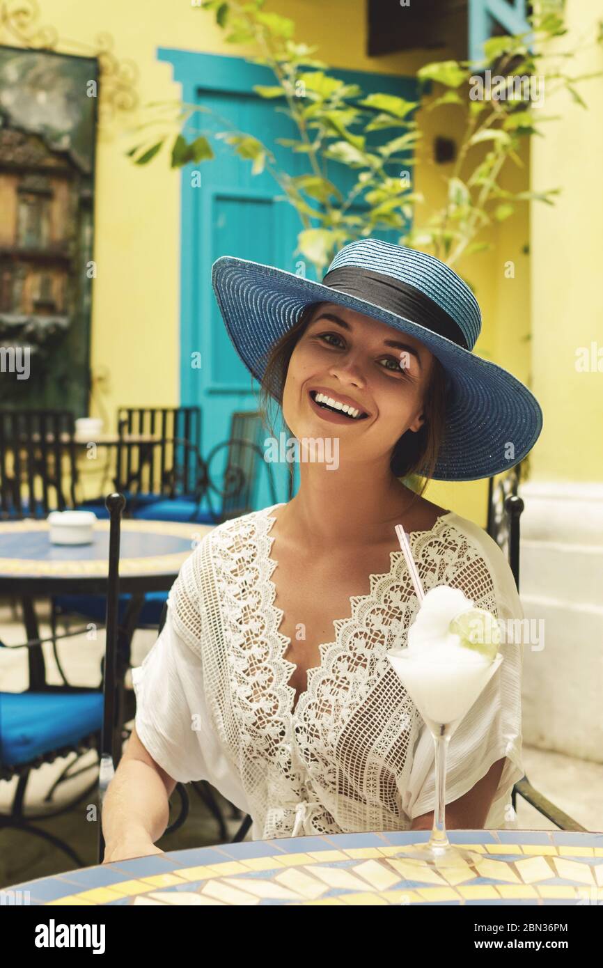 Una donna felice che indossa un cappello blu sta gustando il suo cocktail freddo al bar della città Foto Stock