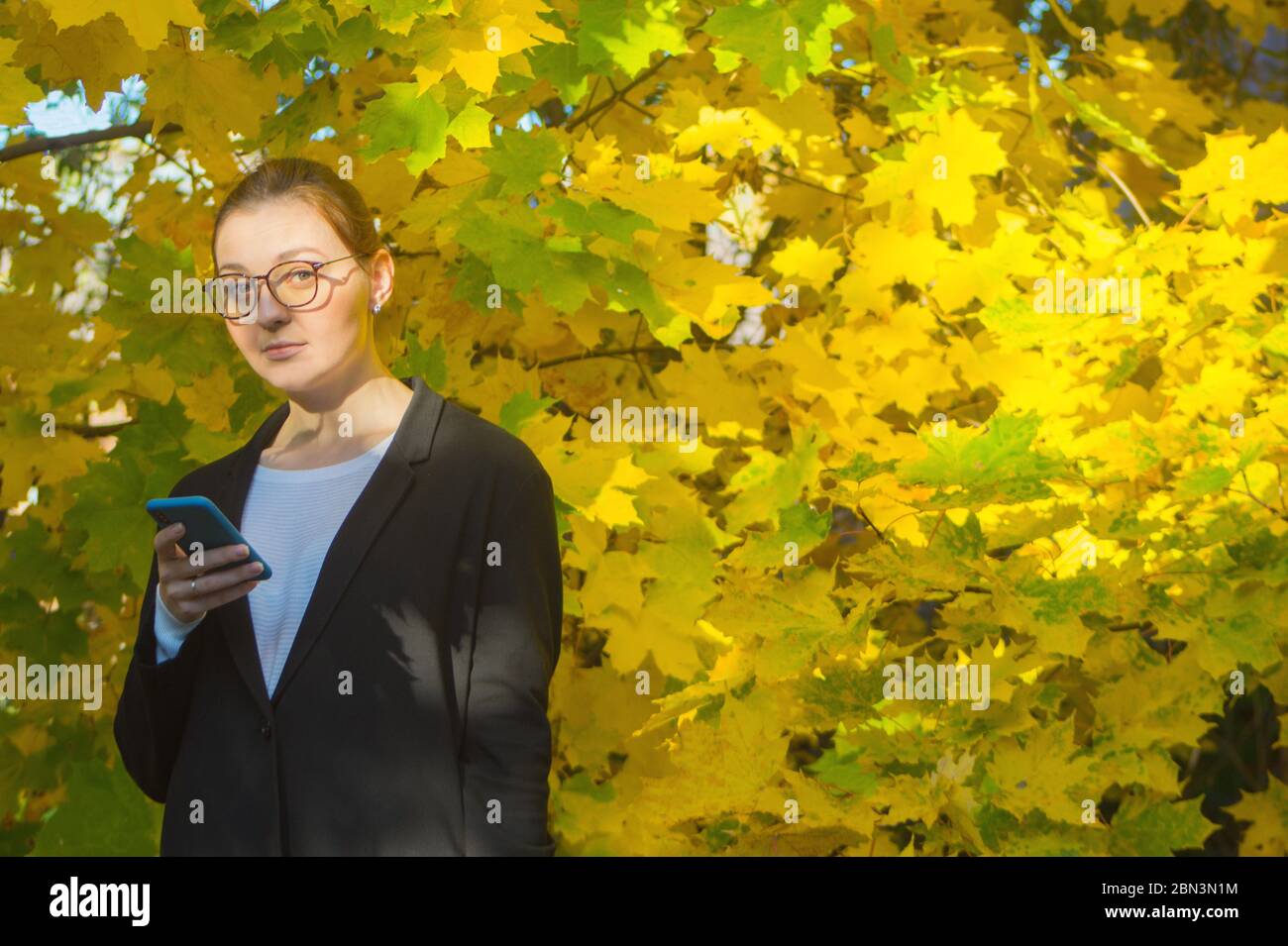 In un caldo giorno d'autunno, una donna guarda leggermente sorpresa alla fotocamera mentre tiene il suo smartphone al livello del petto. Tranquillo ambiente esterno. Foto Stock