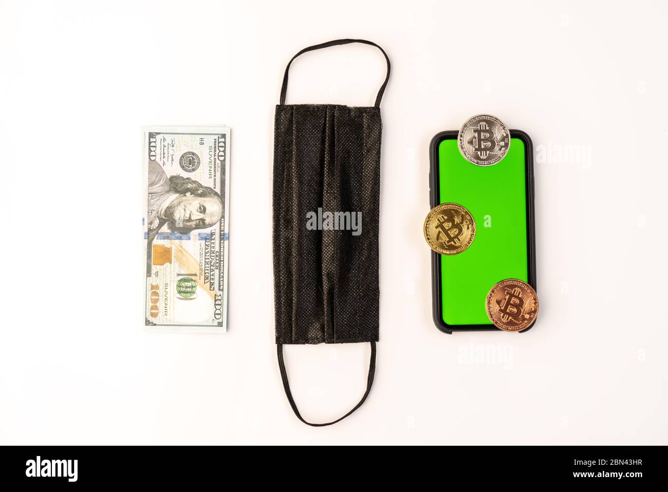 Alcune monete con un segno di bitcoin su un telefono con schermo verde, il dollaro di fattura e maschera protettiva nera Foto Stock