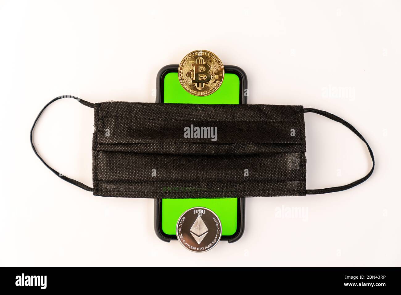 Maschera protettiva nera su un telefono con schermo verde e monete fisiche bitcoin e etereo isolate su sfondo bianco Foto Stock