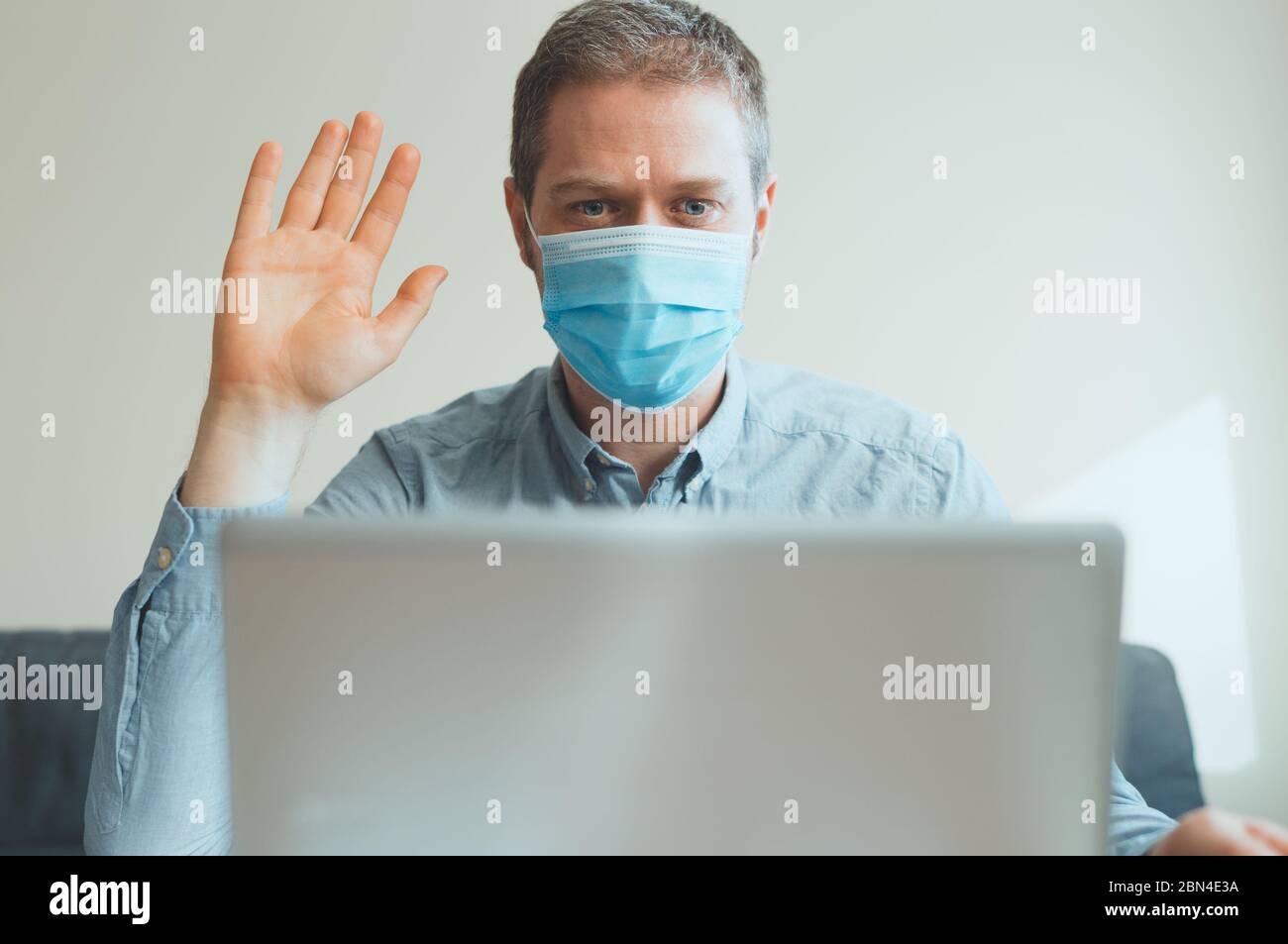 L'uomo in maschera medica saluta via videochiamata in interni. Foto Stock