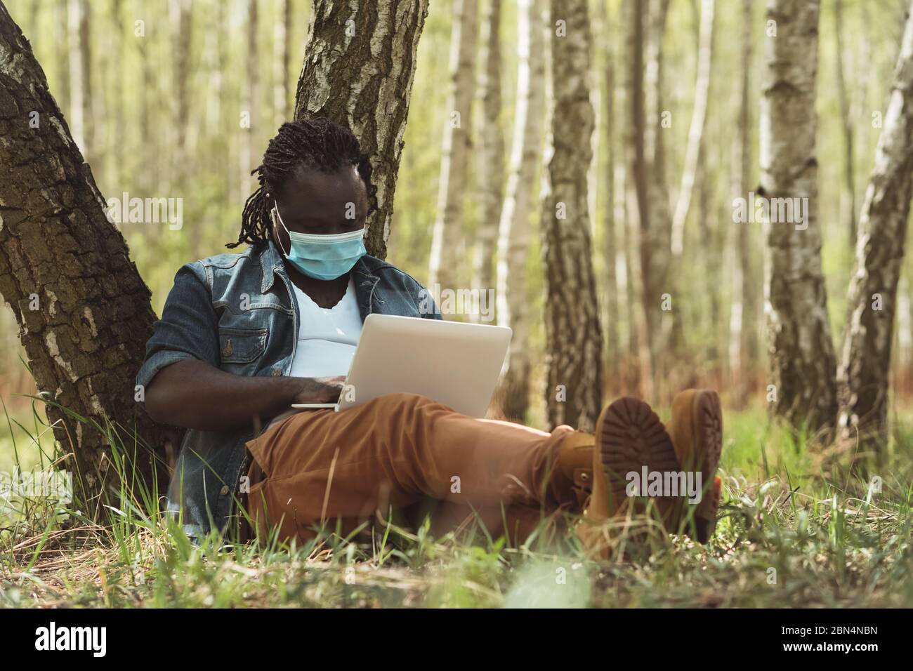 Un uomo africano allegro con maschera medica che fa il lavoro remoto sul laptop nella natura. Focolaio di coronavirus Foto Stock
