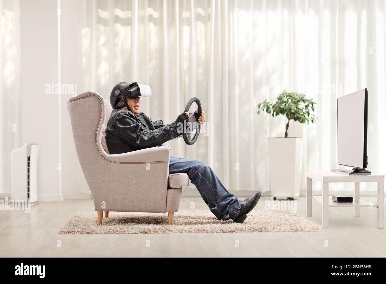 Uomo anziano su una poltrona che utilizza un visore VR e tiene il volante davanti al televisore Foto Stock