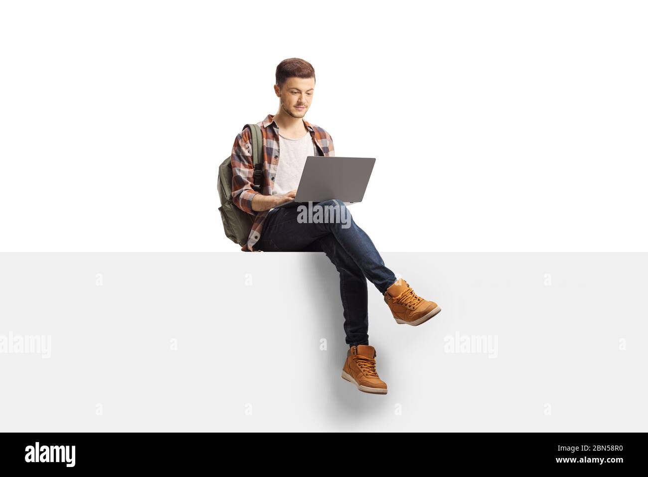 Studente maschio che utilizza un computer portatile e seduto su un pannello vuoto isolato su sfondo bianco Foto Stock