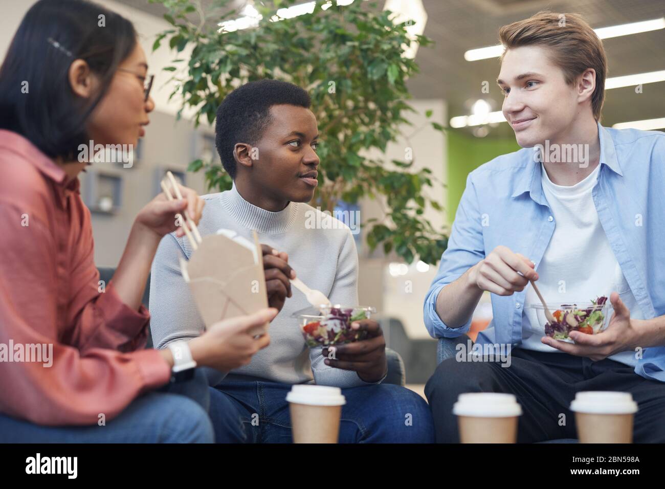 Gruppo multietnico di giovani che mangiano cibo da asporto e sorridono durante la pausa pranzo a scuola o in ufficio Foto Stock