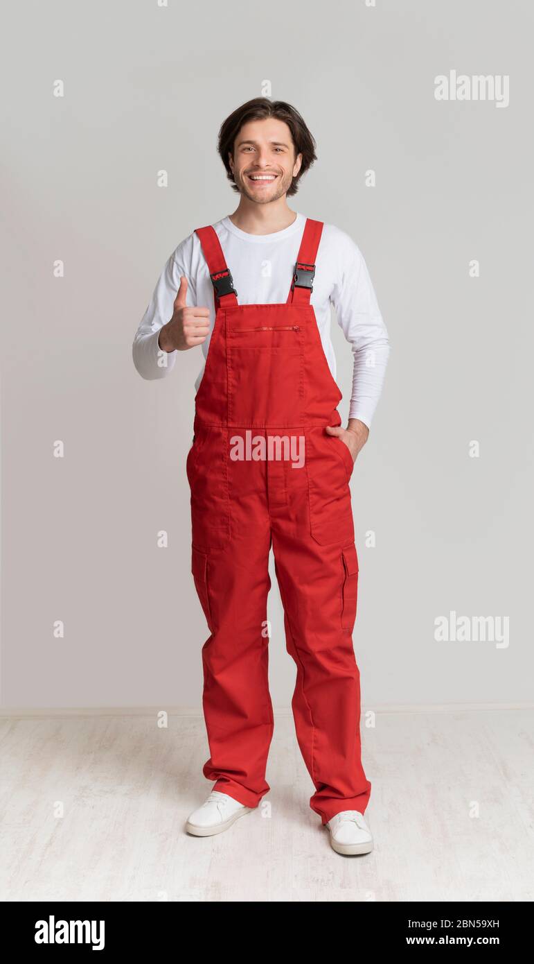 Sorridente giovane operaio che posa in tuta da lavoro e mostra pollice in su Foto Stock