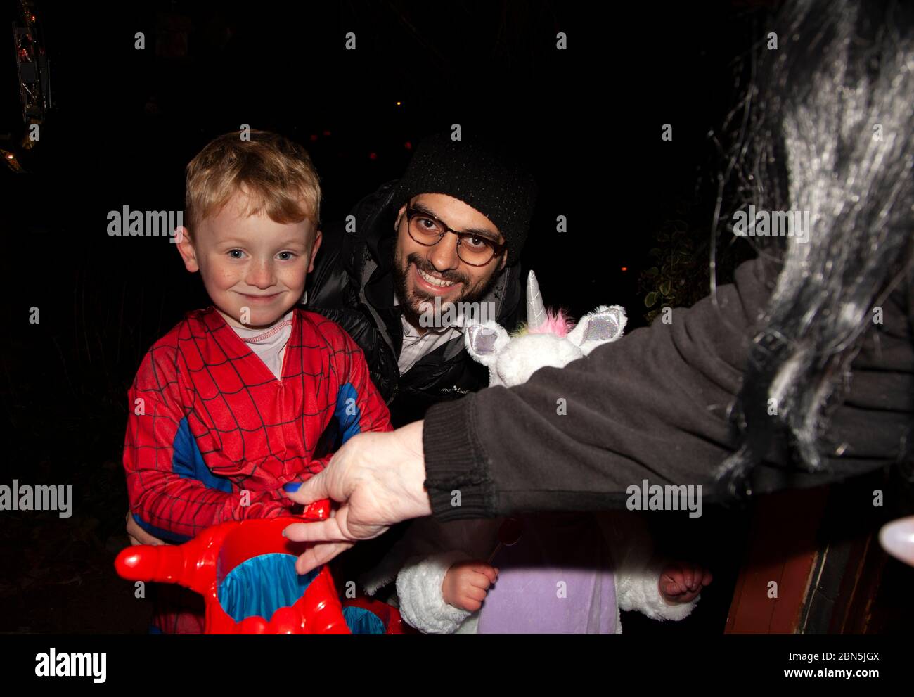 I bambini in costumi Spiderman e Unicorn con il loro papà godendosi Halloween ottenere le delizie da una strega alla porta di ingresso. St Paul Minnesota, Minnesota, USA Foto Stock