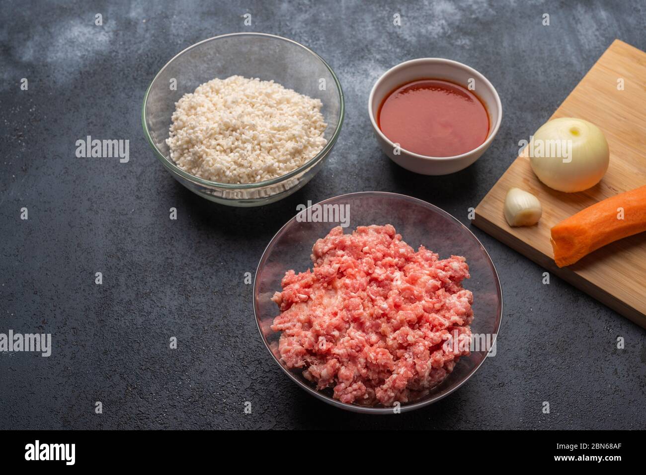Ingredienti per polpette - carne tritata, riso, pasta di pomodoro, cipolla, aglio, carote su un tagliere su sfondo scuro Foto Stock