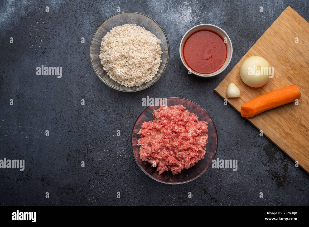 Ingredienti per polpette - carne tritata, riso, pasta di pomodoro, cipolla, aglio, carote su un tagliere su sfondo scuro. Vista dall'alto Foto Stock