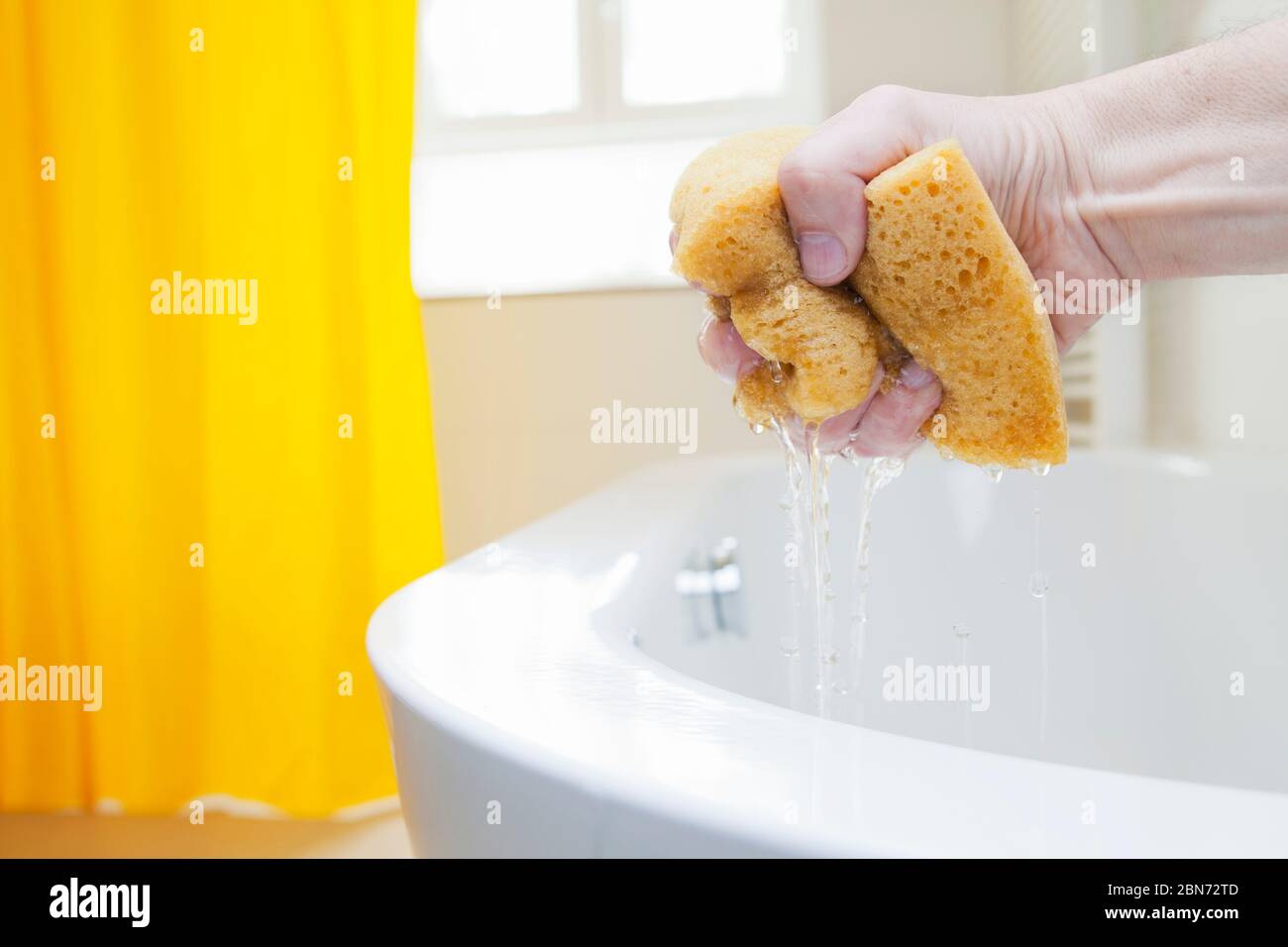 Mano che stringe una spugna con acqua in un bagno luminoso - fuoco sulle gocce d'acqua Foto Stock