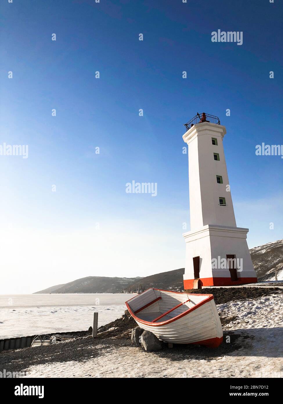 Foto del vecchio faro e della barca di legno sulla riva di Magadan, Russia. Alto edificio bianco e barca a strisce rosse. Cielo azzurro chiaro e ghiaccio sul mare in inverno. Spazio di copia per il testo Foto Stock