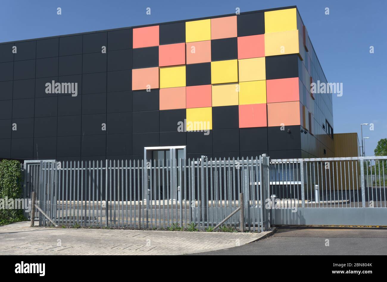 Edificio cubico nero opaco con piastrelle gialle e rosse in un angolo come design grafico dietro le ringhiere di sicurezza in metallo, data center cielo blu intenso Foto Stock
