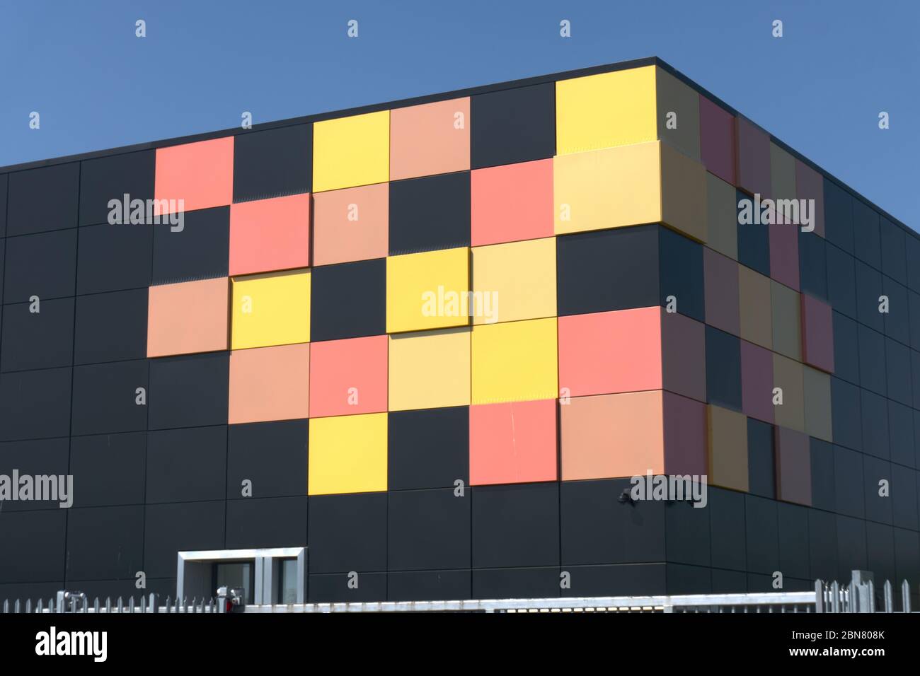 Edificio cubico nero opaco con piastrelle gialle e rosse in un angolo come design grafico dietro le ringhiere di sicurezza in metallo, data center cielo blu intenso Foto Stock