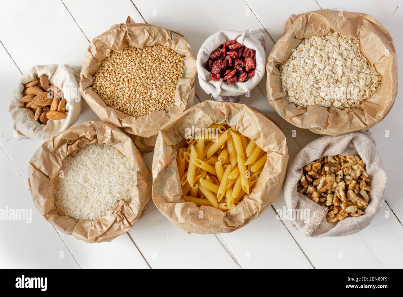 Semole, pasta, noci e frutta secca in sacchetti di carta e cotone su tavola di legno bianco. Vista dall'alto. Zero sprechi, eco-compatibile o concetto di stile di vita senza plastica. Foto Stock