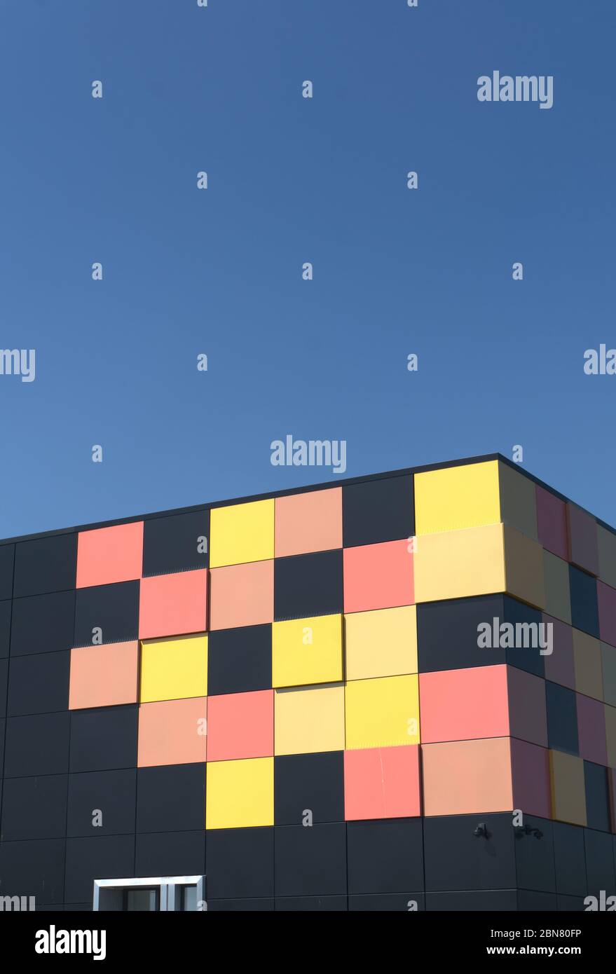Edificio cubico nero opaco con piastrelle gialle e rosse in un angolo come design grafico dietro le ringhiere di sicurezza in metallo, data center cielo blu intenso Foto Stock