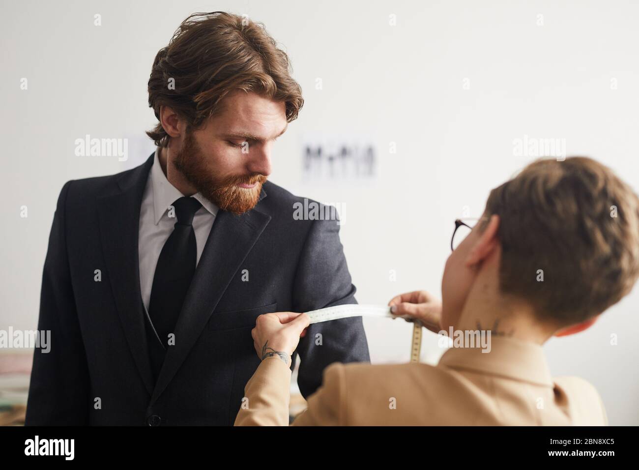 Giovane uomo con il bearded che indossa la tuta mentre misura su misura con il metro a nastro in officina Foto Stock
