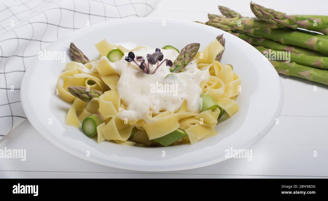 Piatto di pasta fresca fatta in casa di fettuccine o tagliatelle, asparagi verdi, salsa bianca, in un piatto bianco su tavola di fondo bianco in legno Foto Stock