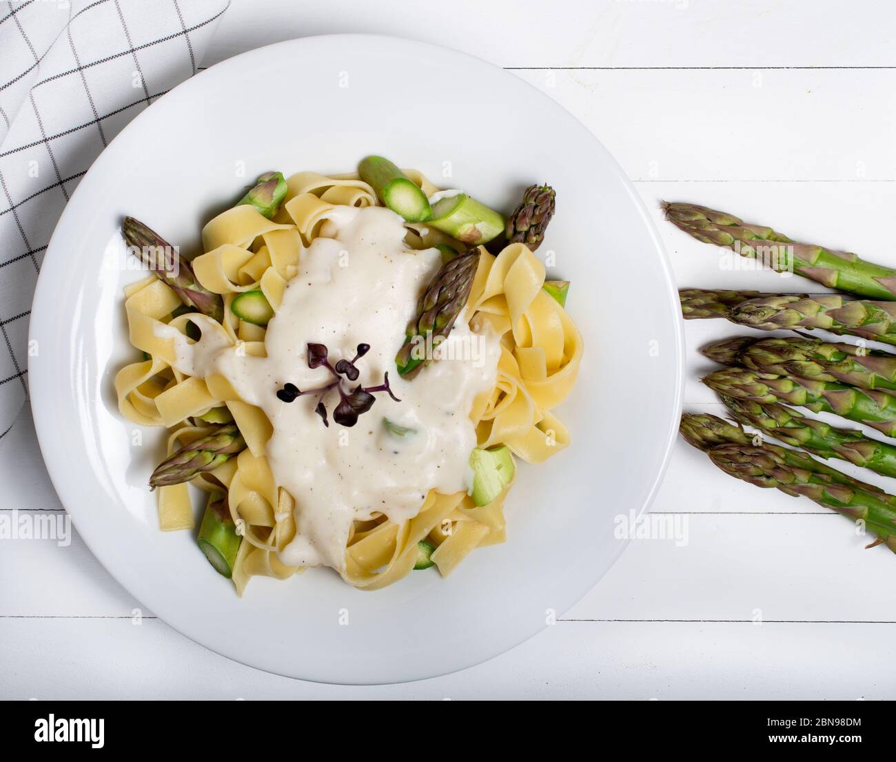 Piatto di pasta fresca fatta in casa di fettuccine o tagliatelle, asparagi verdi, salsa bianca, in un piatto bianco su tavola di fondo bianco in legno Foto Stock