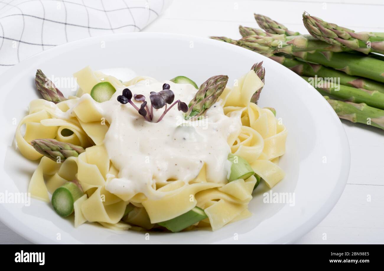 Piatto di pasta fresca fatta in casa di fettuccine o tagliatelle, asparagi verdi, salsa bianca, in un piatto bianco su tavola di fondo bianco in legno Foto Stock