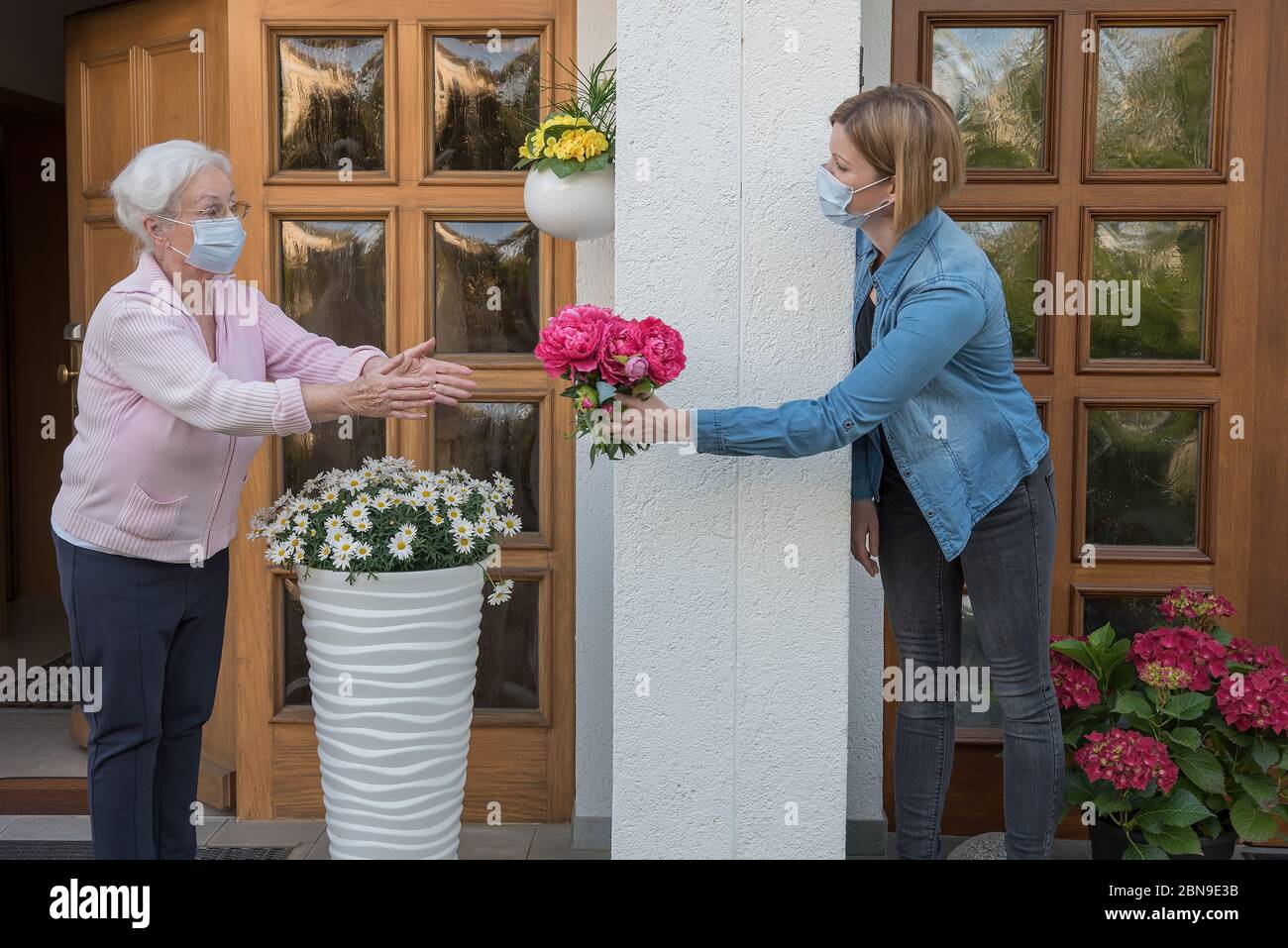Donna anziana con maschera facciale ottiene fiori dalla donna vicina alla porta della casa Foto Stock
