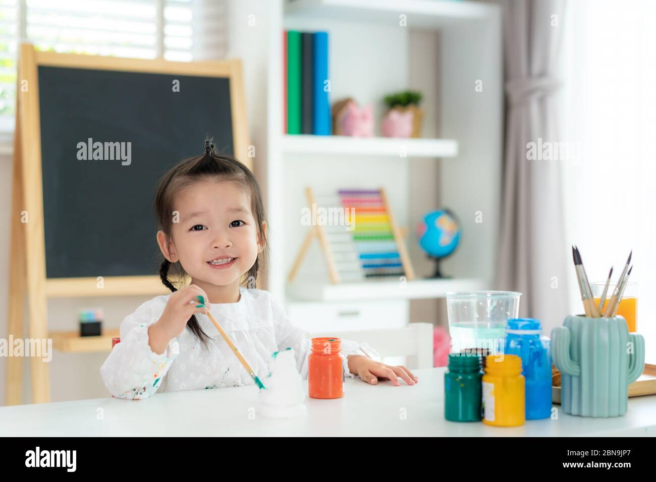 Ragazza asiatica scuola asilo guardando la macchina fotografica e sorridere mentre dipingendo la bambola di Plaster con vernice acrilica di colore dell'acqua nel soggiorno a casa. Homescho Foto Stock