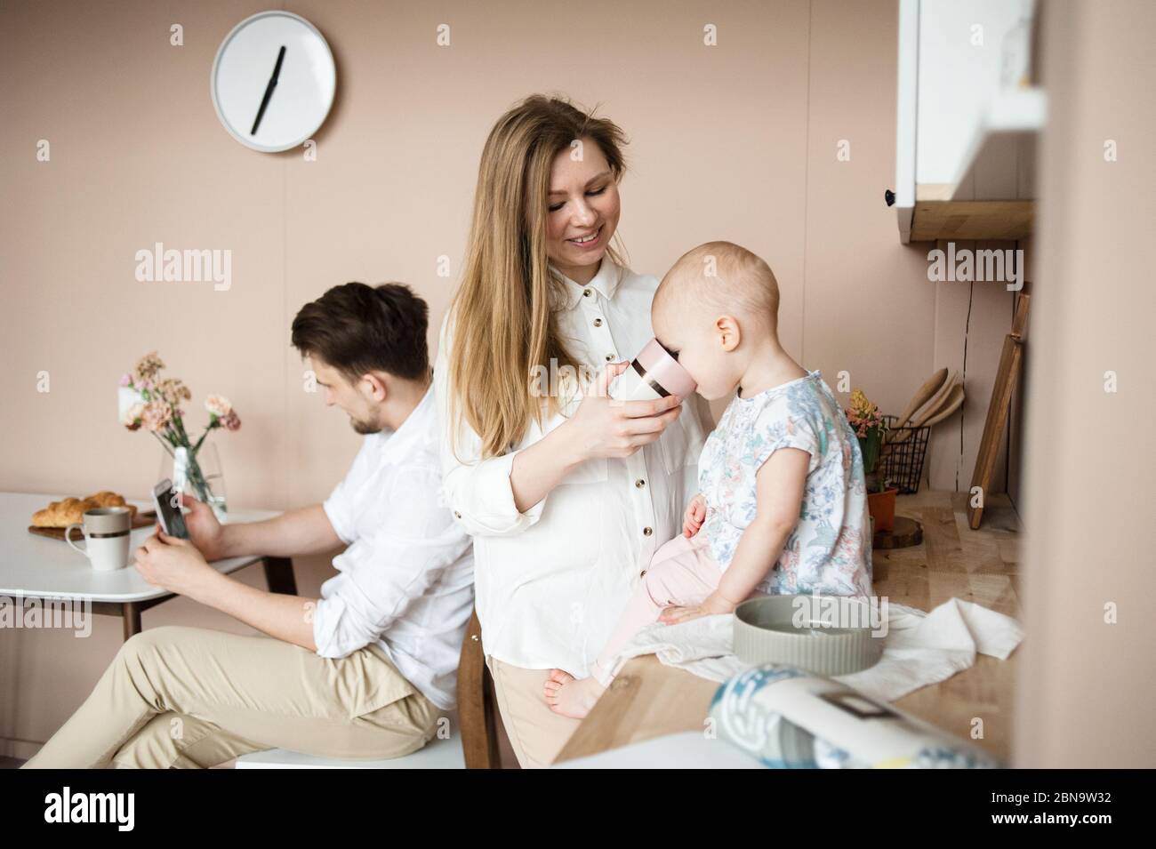 la madre alimenta il bambino seduto sul tavolo della cucina mentre il padre chiacchiera in linea. Foto Stock