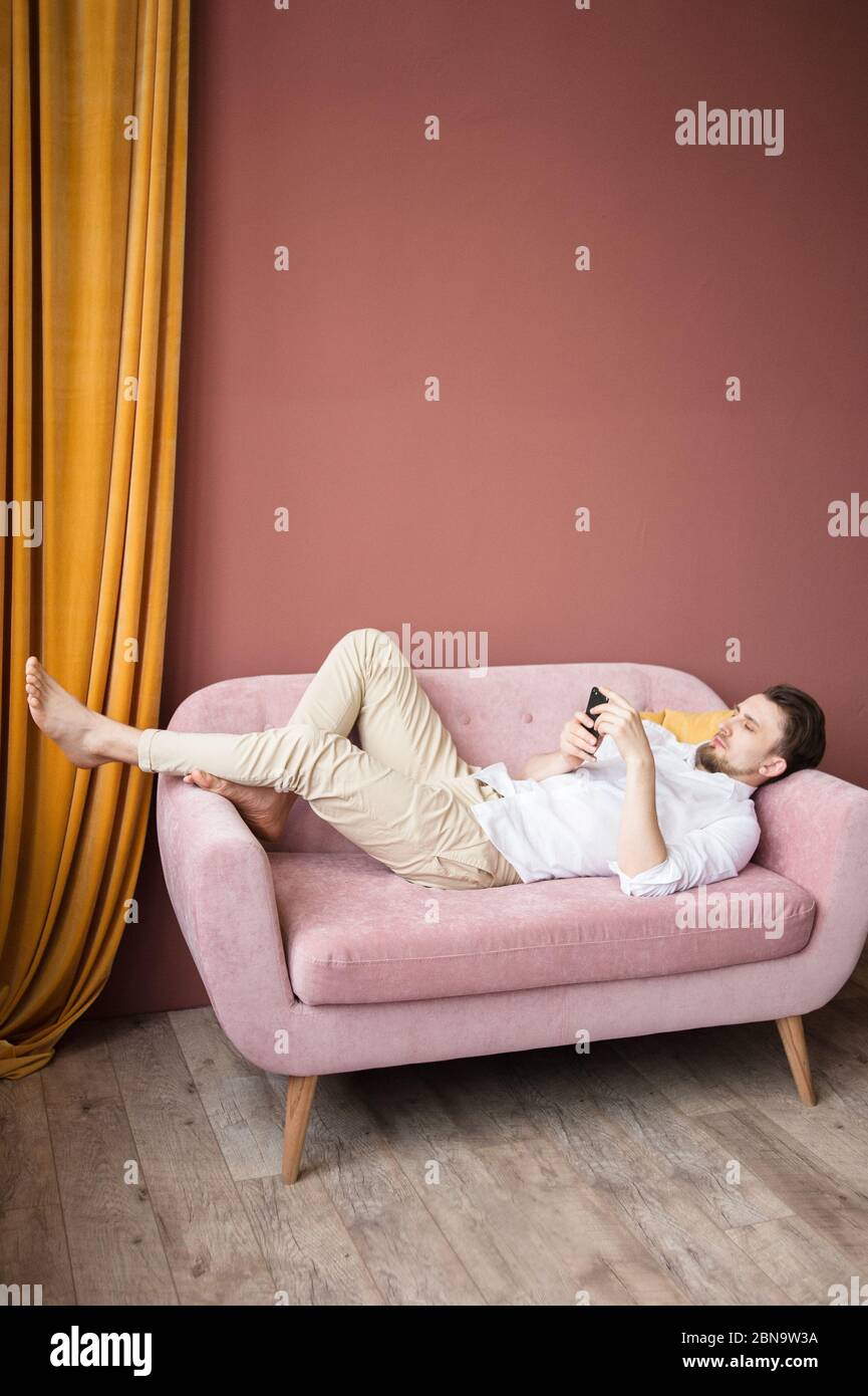 Il giovane in una camicia bianca lavora sul suo telefono mentre si sdraia su un divano rosa Foto Stock
