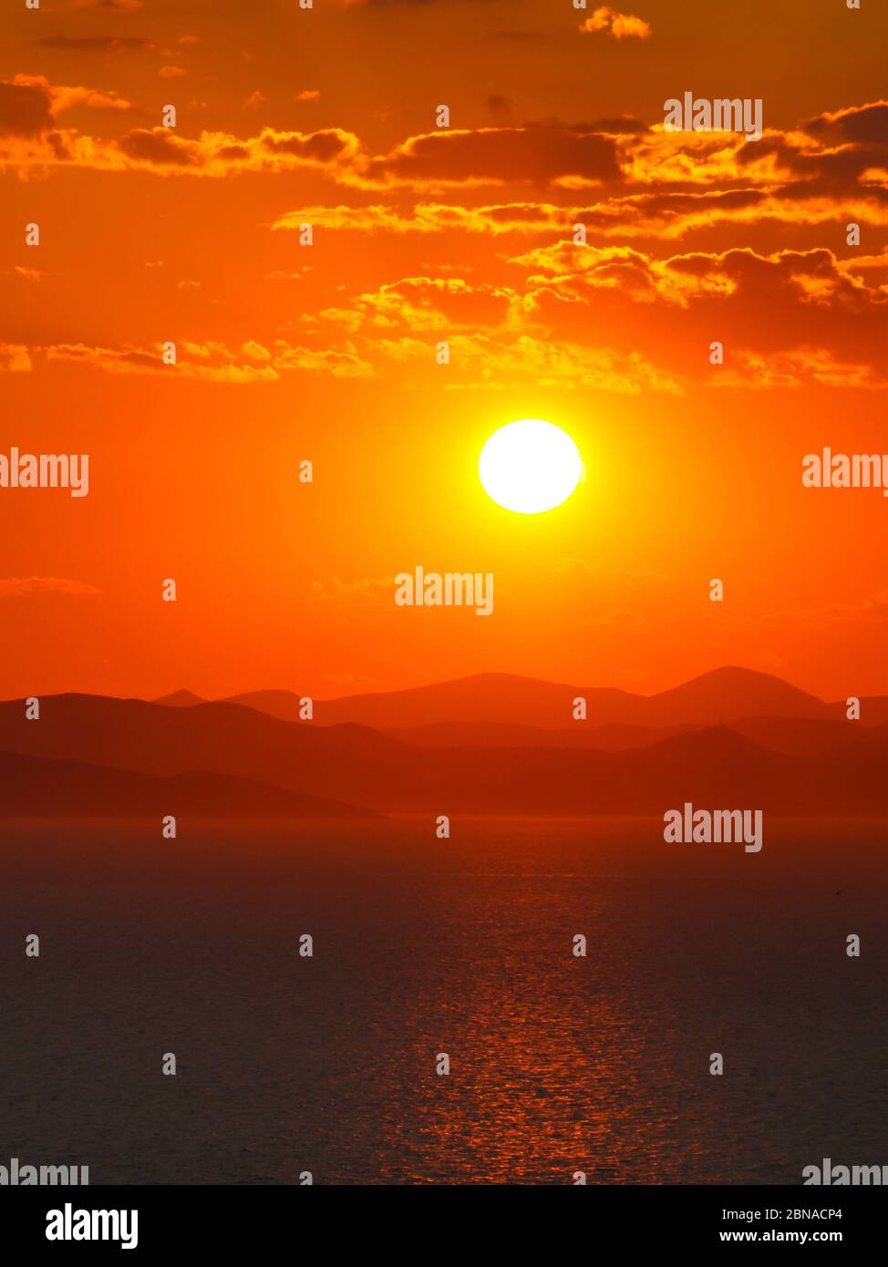 Tramonto luminoso sopra la silhouette delle montagne e il mare. I raggi solari dipingono con il colore arancione lo sfondo dell'orizzonte. Sfondo, foto verticale. Foto Stock