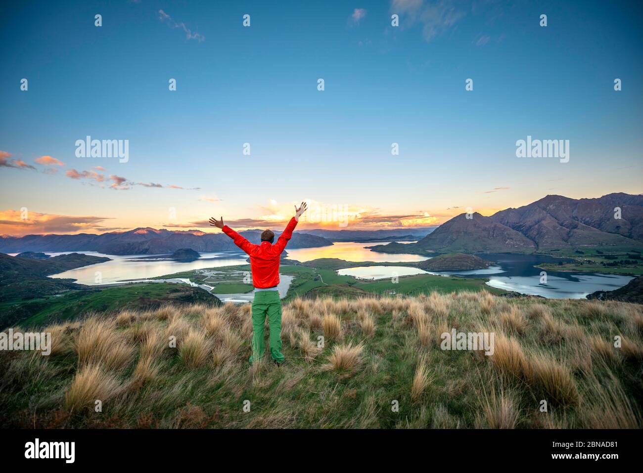 Escursionista allunga le braccia in aria, vista panoramica del lago Wanaka e le montagne al tramonto, Rocky Peak, Glendhu Bay, Otago, South Island Foto Stock