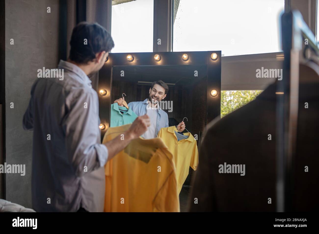 Uomo che riflette nello specchio con le magliette nelle mani Foto Stock