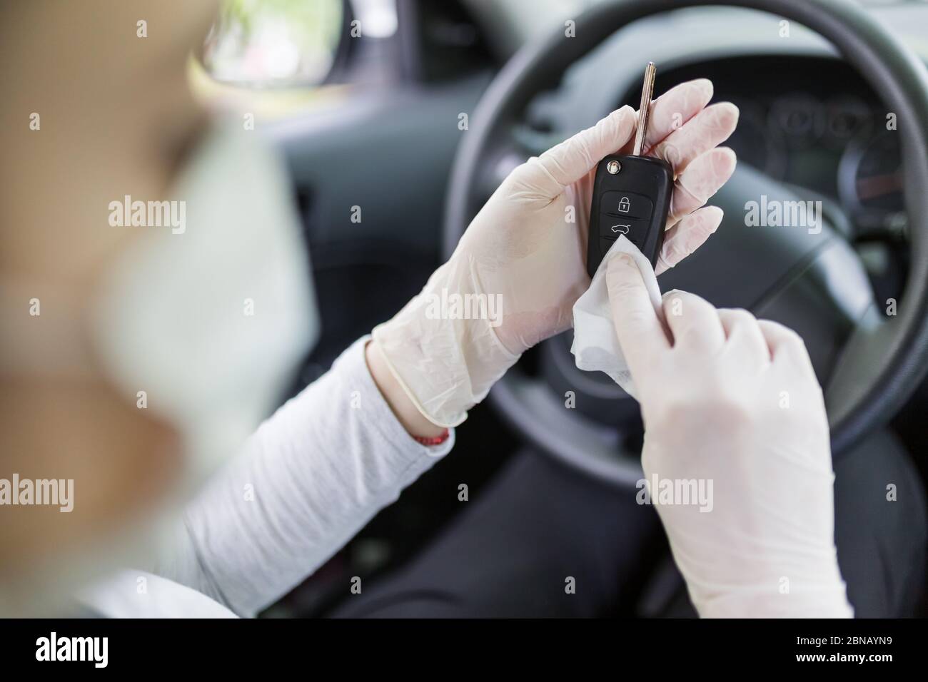 Primo piano della maschera chirurgica protettiva da indossare da donna e indossare guanti in gomma disinfettando la chiave dell'auto con un panno umido all'interno del veicolo. Foto Stock