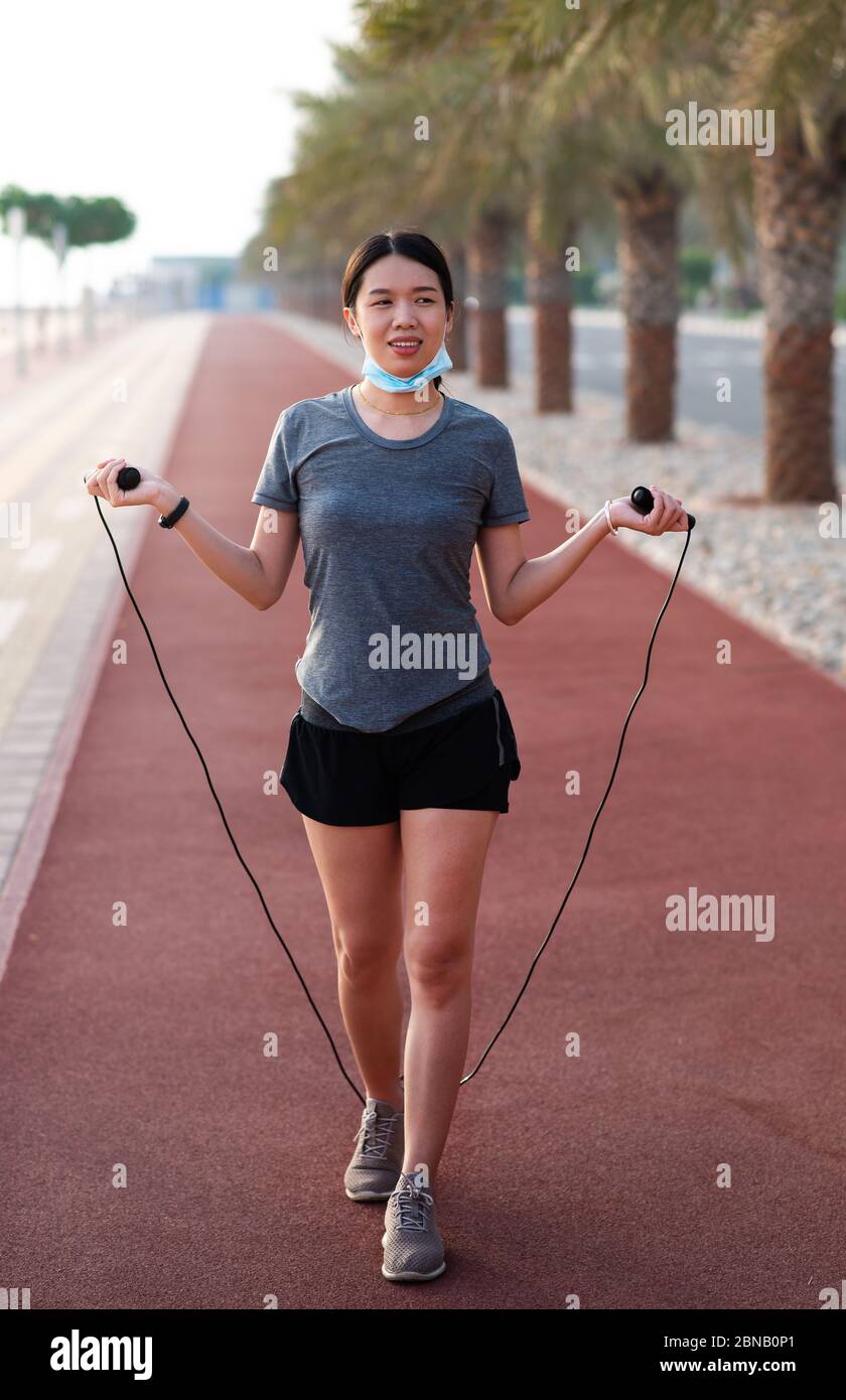 Asian Donna esercizio con una corda di salto con maschera chirurgica protettiva abbassata all'aperto Foto Stock