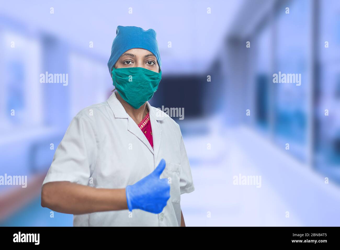 Ritratto di medico lavoratore femminile medico medico indossando maschera chirurgica e cappuccio facendo Thumbs Up, OK segno, eccellente segno. Corridoio esterno dell'ospedale. Covid-19 Foto Stock