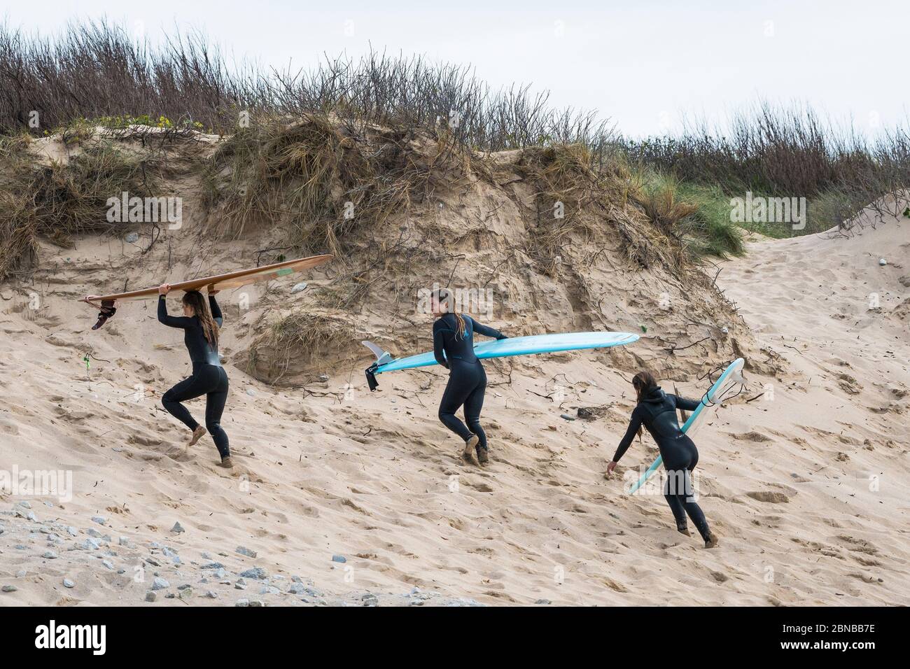 Giovani surfisti femminili che portano le loro tavole da surf e salgono sulle dune di sabbia dopo il surf a Fistral a Newquay in Cornovaglia. Foto Stock