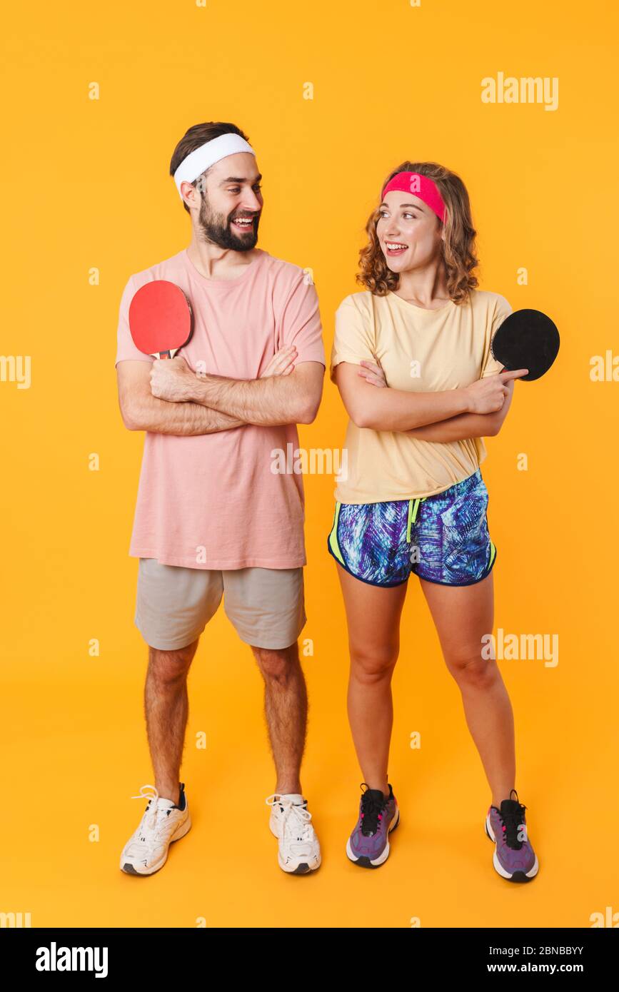 Ritratto di giovane coppia sportiva felice con archetti che sorridono e tengono racchette da ping pong isolate su sfondo giallo Foto Stock